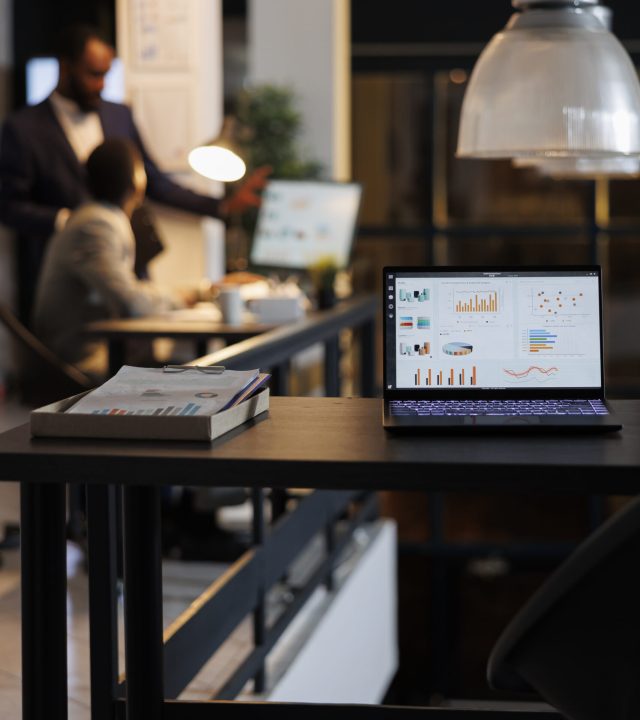 Laptop computer with financial graphs on screen, standing on table in startup office, coworkers discussing company strategy in background. Employees working late at night in startup office